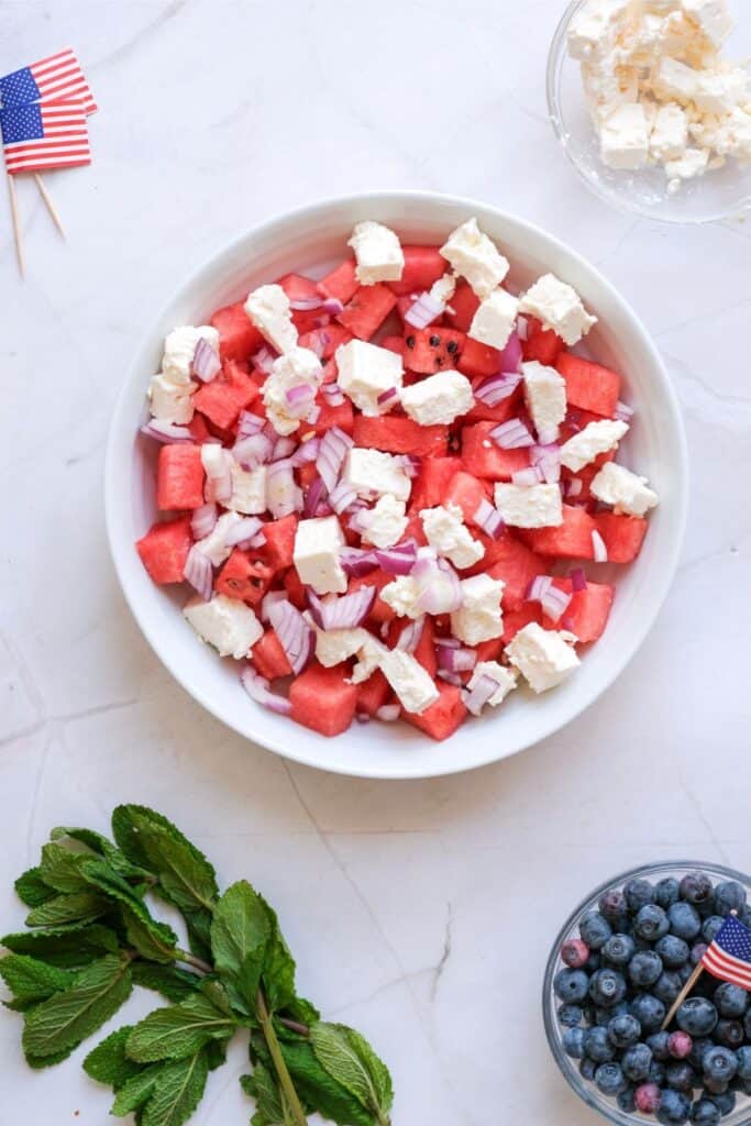 A bowl of Watermelon Feta Salad with diced red onion sits on a white surface near mint leaves, blueberries, crumbled feta, and small American flags.