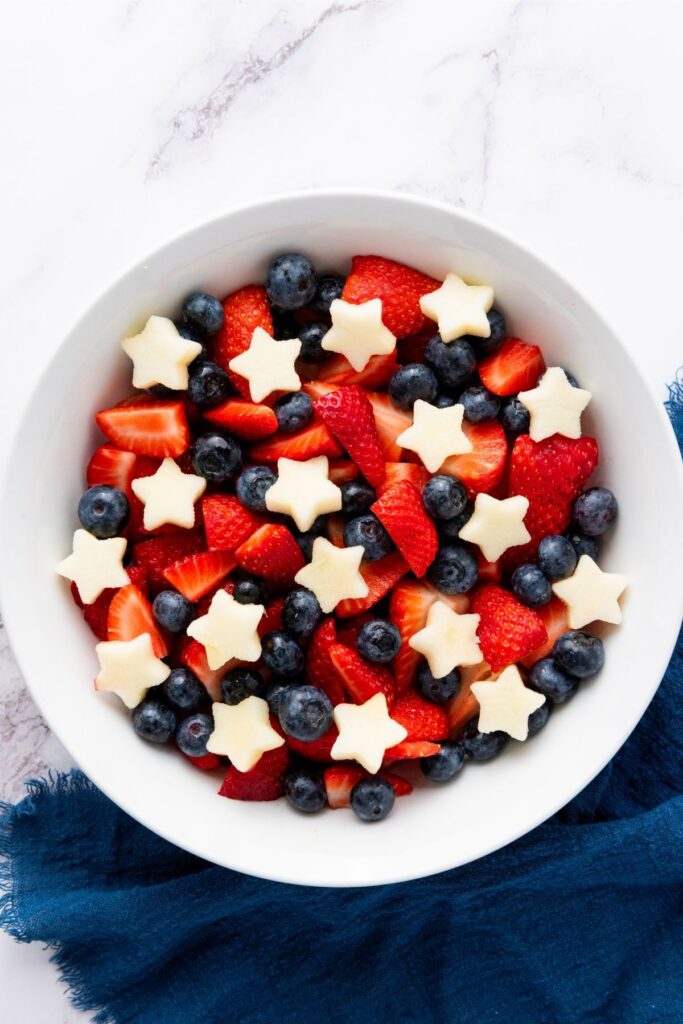 A white bowl filled with strawberries, blueberries, and star-shaped pieces of fruit creates a festive Red White Blue Fruit Salad, placed on a blue cloth on a marble surface.