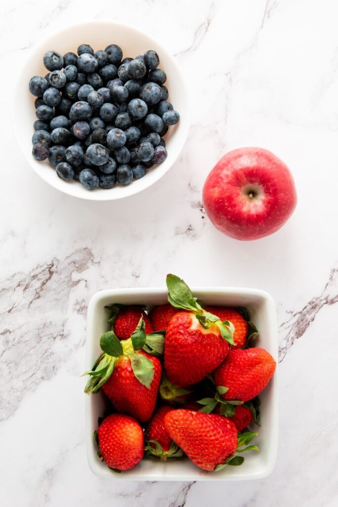 A white bowl of blueberries, a red apple, and a square white bowl of strawberries on a white marble surface—perfect ingredients for a Red White Blue Fruit Salad.