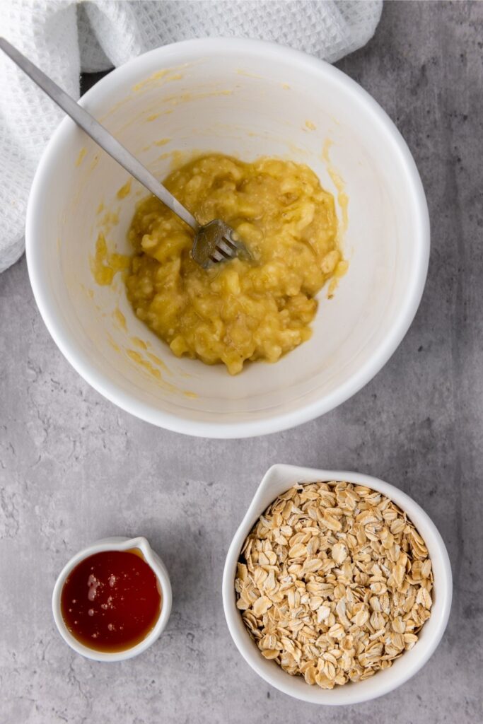 A white bowl with mashed banana and a fork sits next to a small jug of honey and a bowl of rolled oats, perfect for adding to mixed berry granola cups or topping your favorite berry granola on a gray surface.