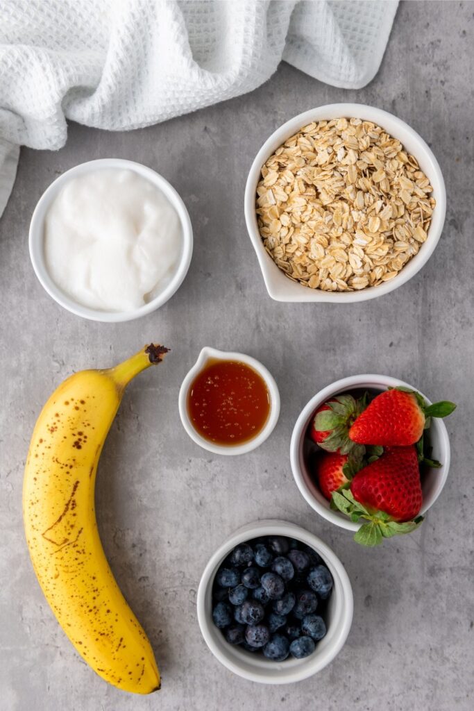 A banana, a bowl of oats, a bowl of strawberries, blueberries, yogurt, honey, and Berry Granola are artfully arranged on a gray surface.