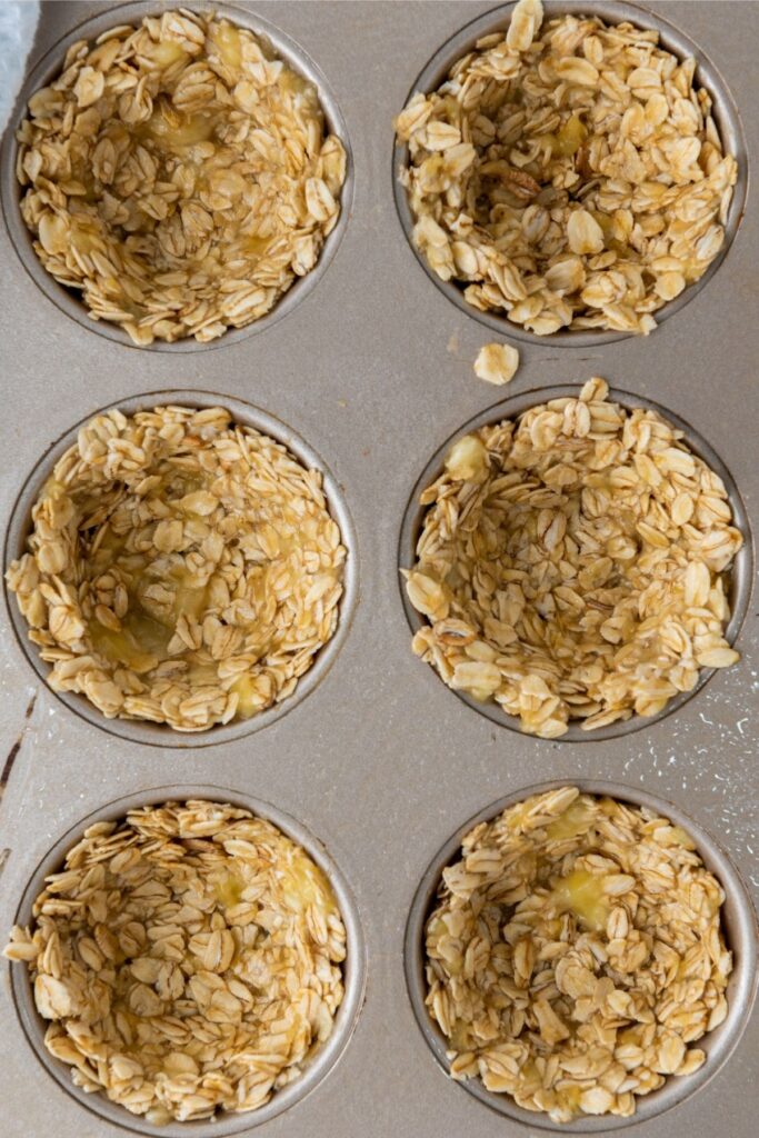 A muffin tin holds six wells filled with a mixture of oats and a yellowish binder, shaped into cups and ready to be baked into delicious Mixed Berry Granola Cups.