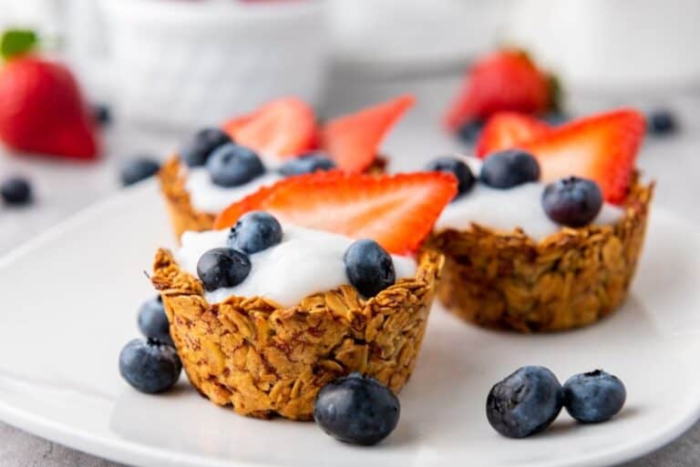 Three granola cups filled with yogurt, topped with sliced strawberries and blueberries, are arranged on a white plate with extra mixed berry scattered around.