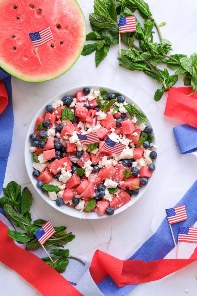A festive watermelon feta salad featuring blueberries, mint, and small American flags, surrounded by red and blue ribbons, mint sprigs, and a halved watermelon.