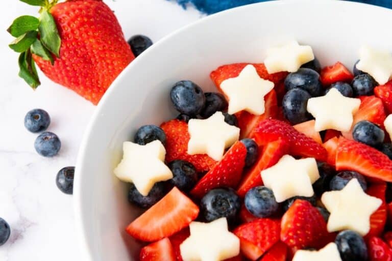 A white bowl filled with sliced strawberries, whole blueberries, and star-shaped pieces of melon creates a vibrant patriotic fruit salad, with a large strawberry and scattered blueberries beside the bowl.
