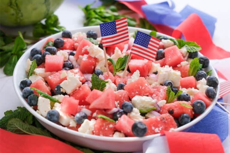 A festive Watermelon Salad featuring watermelon, blueberries, feta cheese, and mint leaves is topped with American flags and surrounded by patriotic red, white, and blue ribbons.