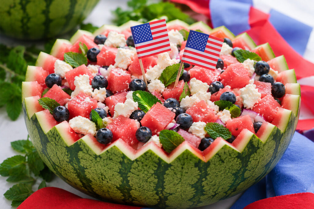 A festive Watermelon Feta Salad served in a watermelon bowl, filled with watermelon cubes, blueberries, mint leaves, and feta cheese, and decorated with two small American flags.
