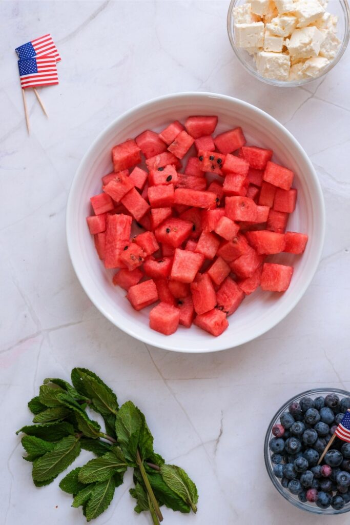 A bowl of cubed watermelon, a bowl of blueberries, a bowl of cubed cheese reminiscent of a classic watermelon feta salad, fresh mint, and two small American flag toothpicks on a marble surface.