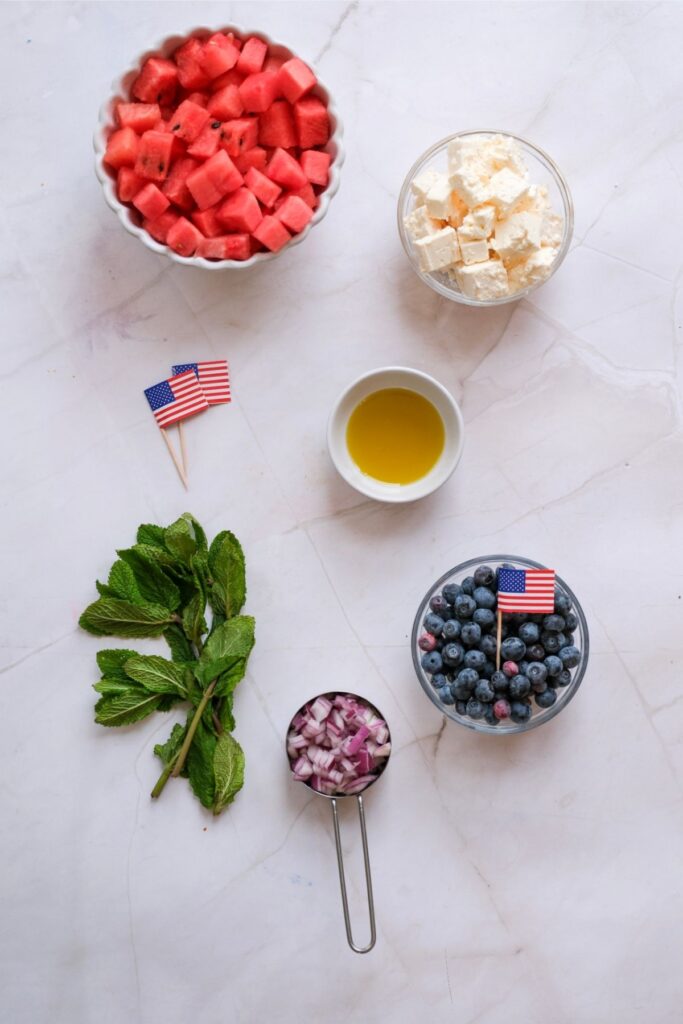 Bowls of Watermelon Feta Salad with blueberries, chopped red onion, fresh mint, and a small dish of olive oil are arranged on a white surface, some topped with tiny American flags.