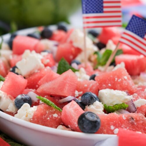 A festive Watermelon Feta Salad featuring a bowl of watermelon, blueberries, feta cheese, and mint, garnished with small American flags; a watermelon half sits in the background.