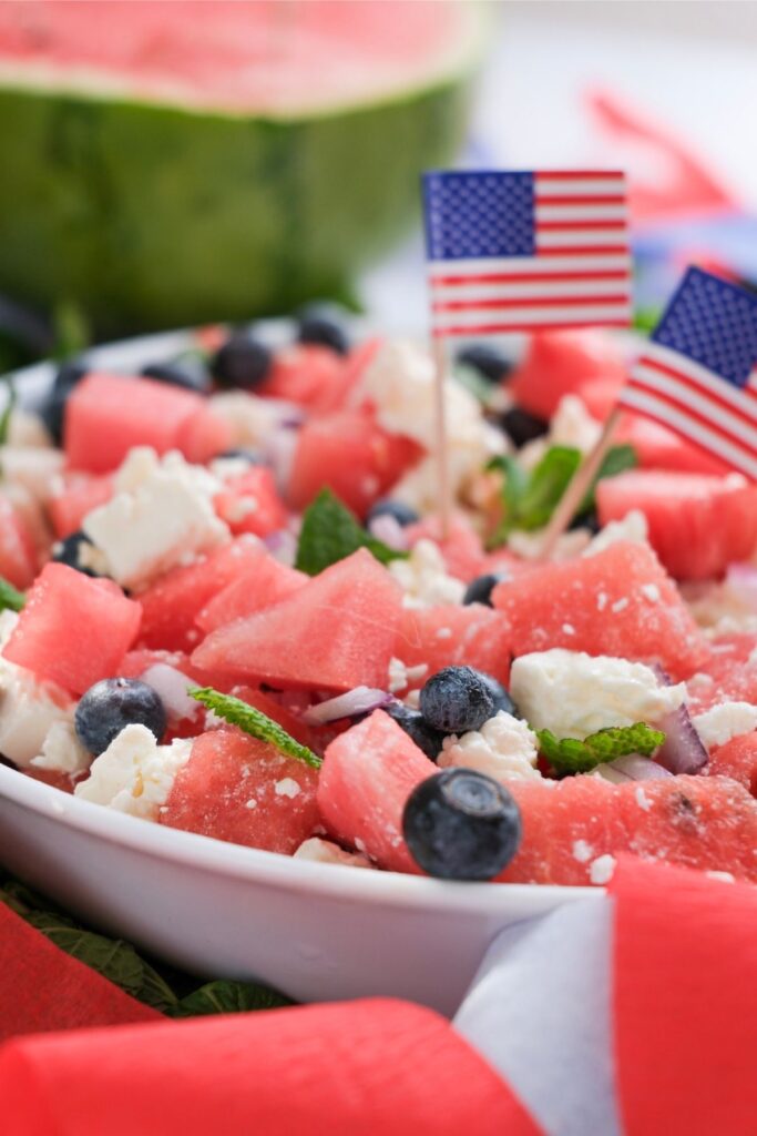 A festive Watermelon Feta Salad featuring a bowl of watermelon, blueberries, feta cheese, and mint, garnished with small American flags; a watermelon half sits in the background.