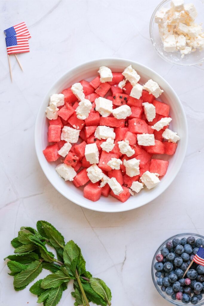 A white bowl filled with cubed watermelon and chunks of feta cheese creates a refreshing Watermelon Feta Salad, surrounded by fresh mint, blueberries, a glass bowl of feta, and small American flag toothpicks on a marble surface.