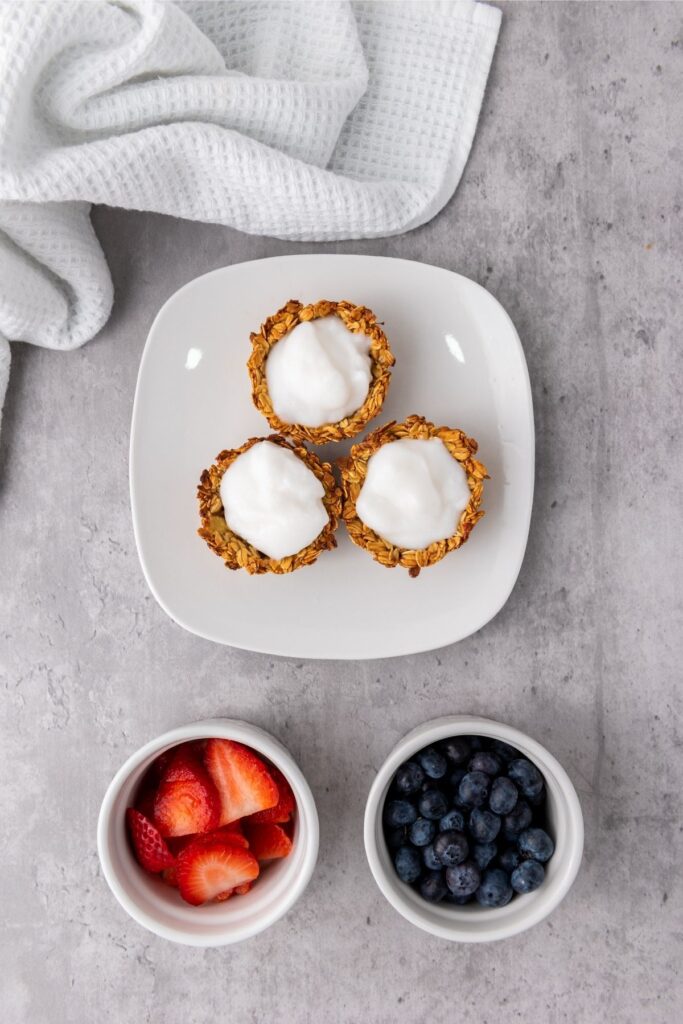 Three Granola Cups with yogurt rest on a white plate, accompanied by small bowls of sliced strawberries and blueberries—a perfect Berry Breakfast—on a gray surface, with a white towel in the corner.