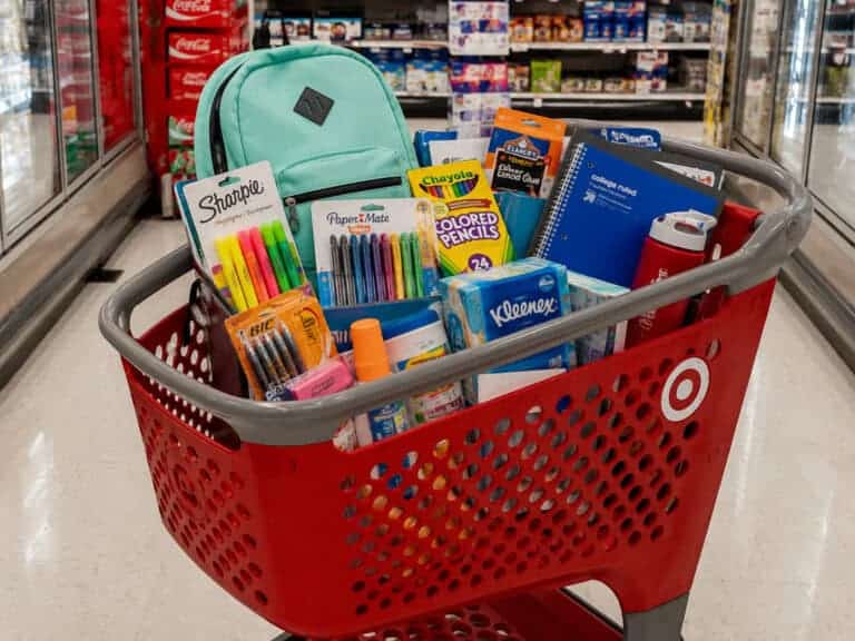 A red Target shopping cart filled with various school supplies, including a mint green backpack, notebooks, pens, markers, colored pencils, tissues, and glue—perfect for college students or teachers stocking up for the new semester.