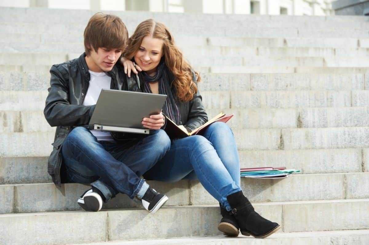 Two young adults, a male and a female, sit on steps looking at a laptop, possibly exploring the latest shows on Amazon Prime or signing up for a free trial together.