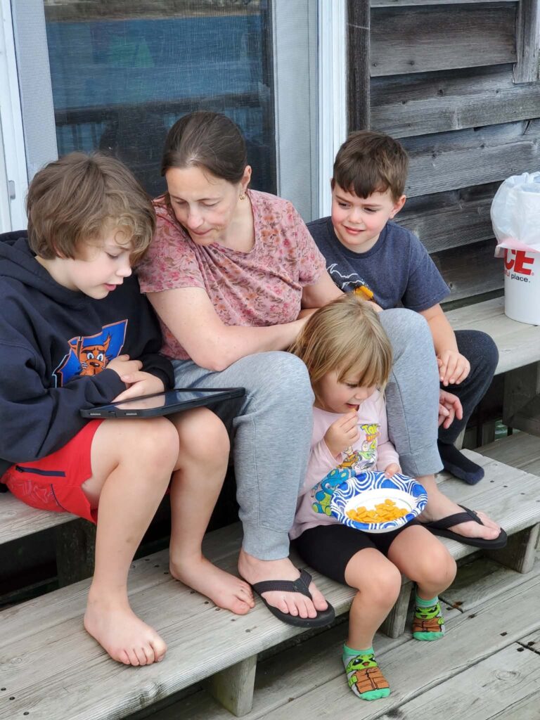 A woman sits on outdoor steps with three children; as one child eats snacks from a bowl and others look at a tablet, it sparks the simple question of what I learned about personal discovery in everyday moments. A plastic bag is in the background.