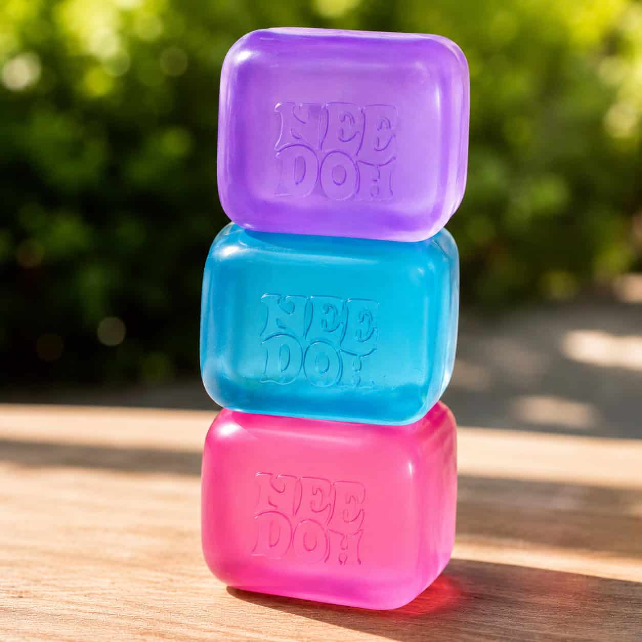 Three rectangular NeeDoh Nice Cube stress balls in purple, blue, and pink are stacked on a wooden surface outdoors, with greenery in the blurred background. Enjoy free style fidget fun wherever you go!.