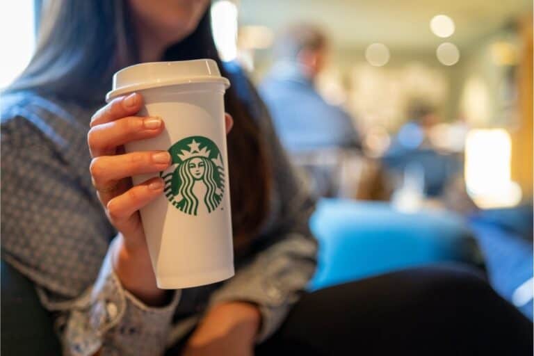 Person holding a white Starbucks coffee cup with a lid; background shows a blurred indoor café setting with people, possibly inside a Target store, capturing the cozy vibe of enjoying Starbucks while shopping.