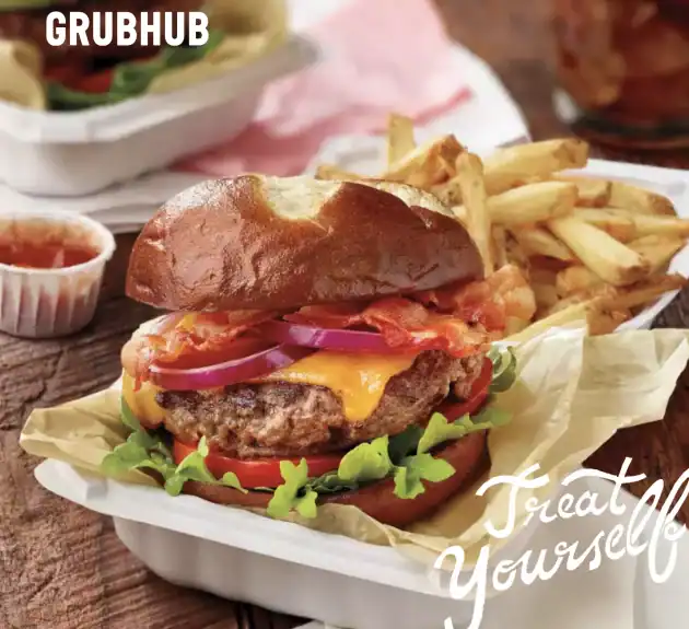 A cheeseburger with lettuce, tomato, bacon, and red onion in a bun, served with French fries and ketchup in a takeout container. Perfect for Amazon Prime young adults. Grubhub logo and "Treat Yourself" text overlay.