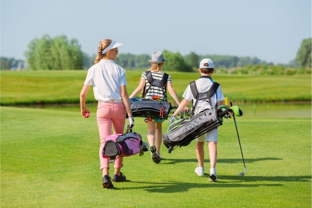Three children carrying golf bags walk across a grassy golf course on a sunny day, enjoying the benefits of kids golf membership and Youth on Course programs.