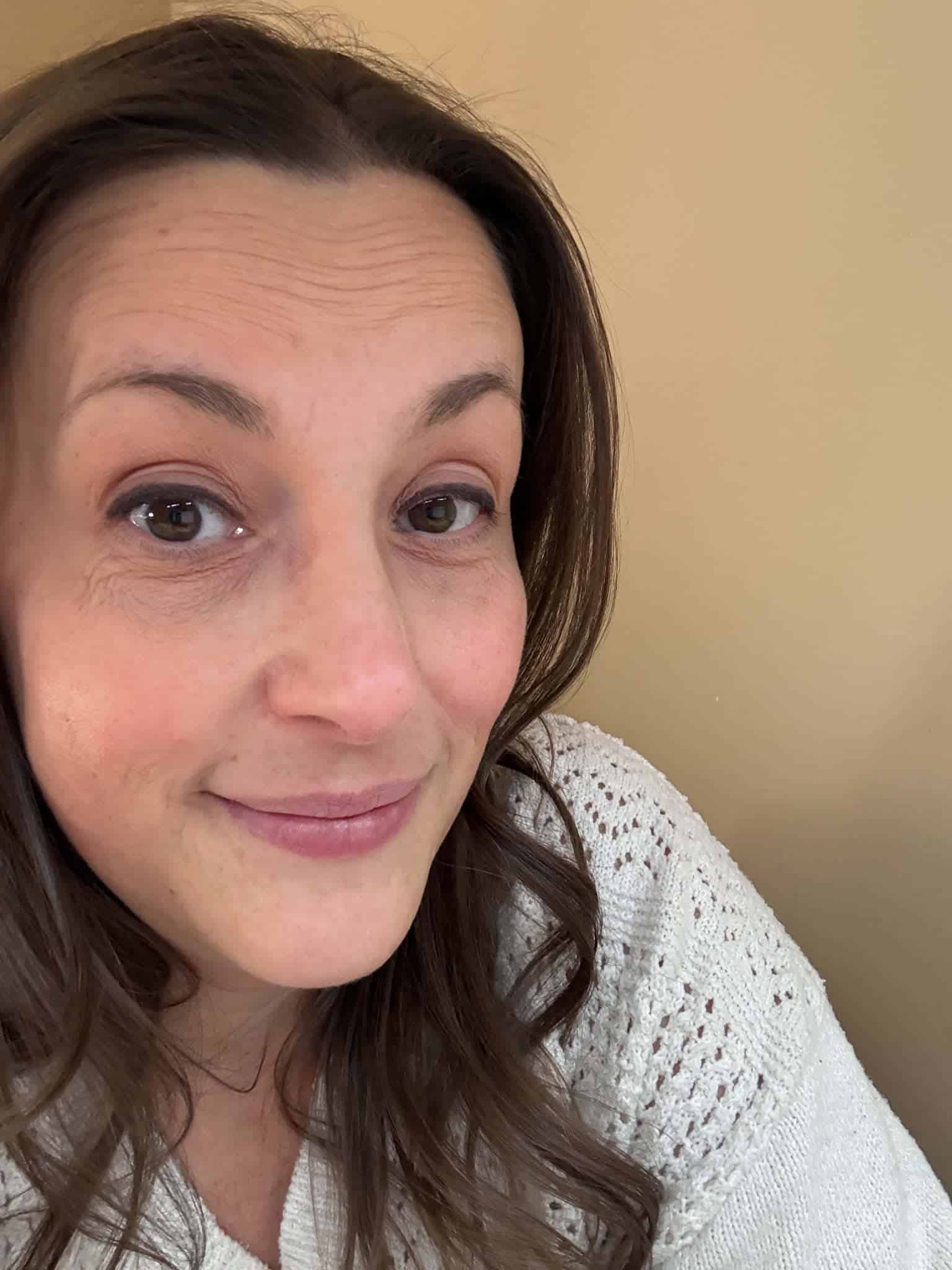 A woman with long brown hair and a white knit sweater smiles slightly while looking at the camera against a beige background, as if pondering what I learned from a simple question.