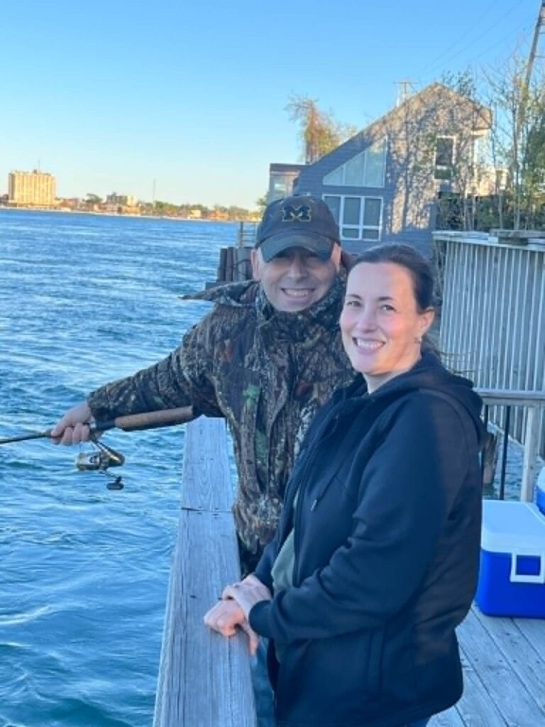 Two people stand by a wooden railing near the water; one holds a fishing rod while the other smiles at the camera, reflecting on lessons learned. A cooler and buildings are visible in the background.