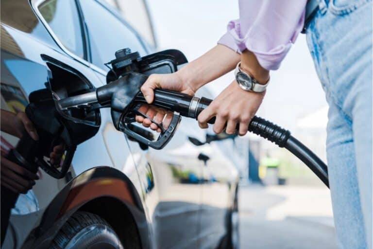 A person refuels a black car at a gas station, nozzle inserted into the open fuel tank.