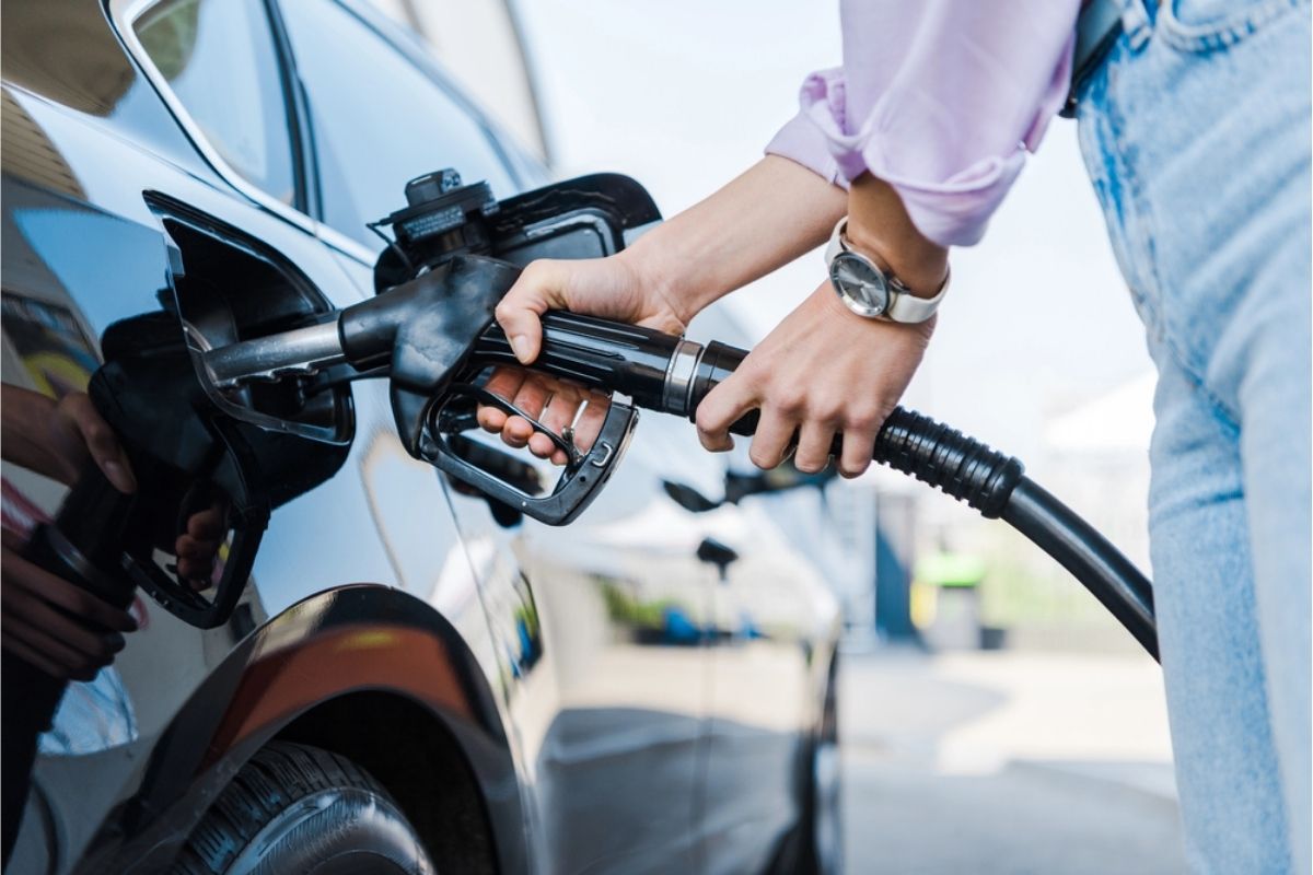 A person refuels a black car at a gas station, nozzle inserted into the open fuel tank.