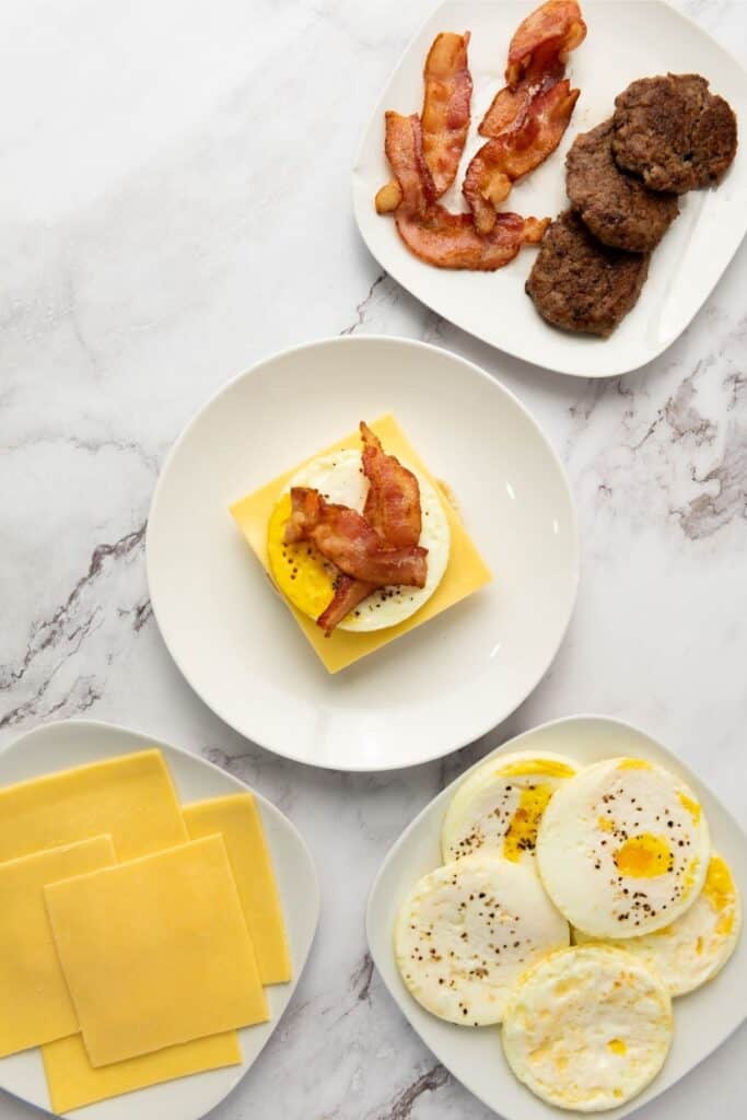 Cooked bacon, sausage patties, fried eggs, cheddar slices, and a breakfast sandwich on a white surface.