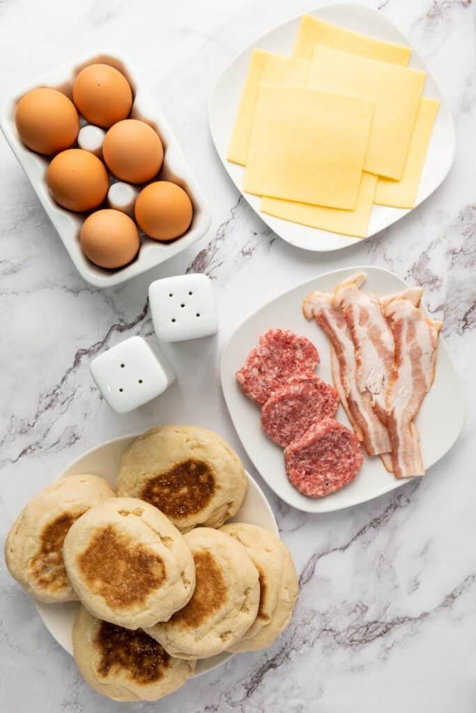 Eggs, cheese slices, sausage patties, bacon, English muffins, and salt and pepper shakers on a marble counter.