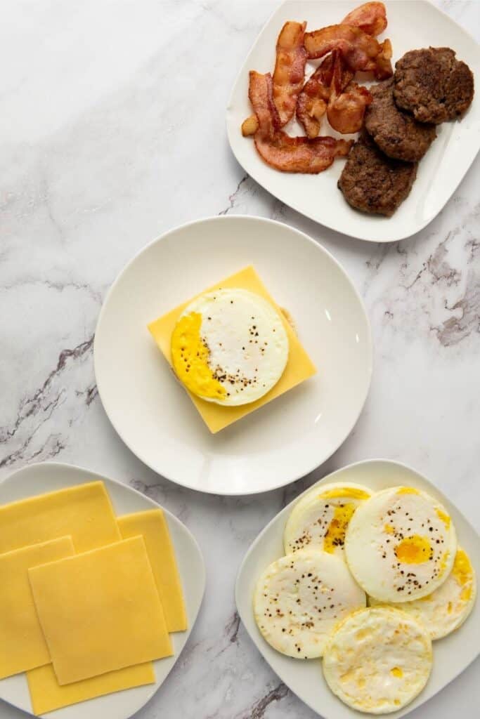 Cooked bacon, sausage patties, cheese slices, and fried eggs neatly arranged on plates atop a marble surface.