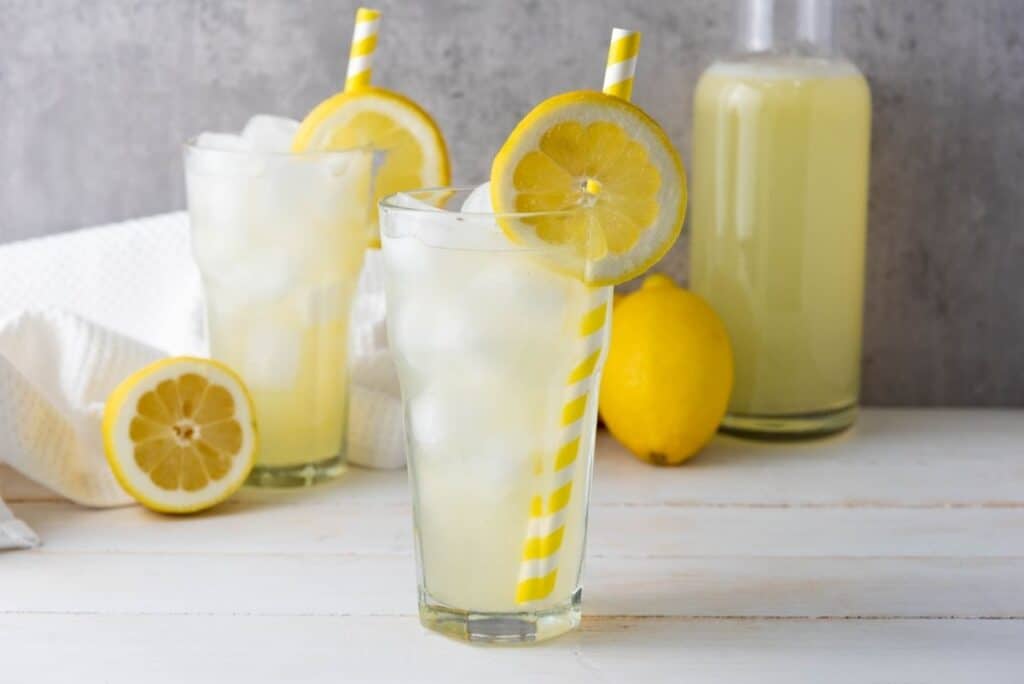 Two glasses of iced lemonade with yellow striped straws, lemon slices, a whole lemon, and a bottle on white.
