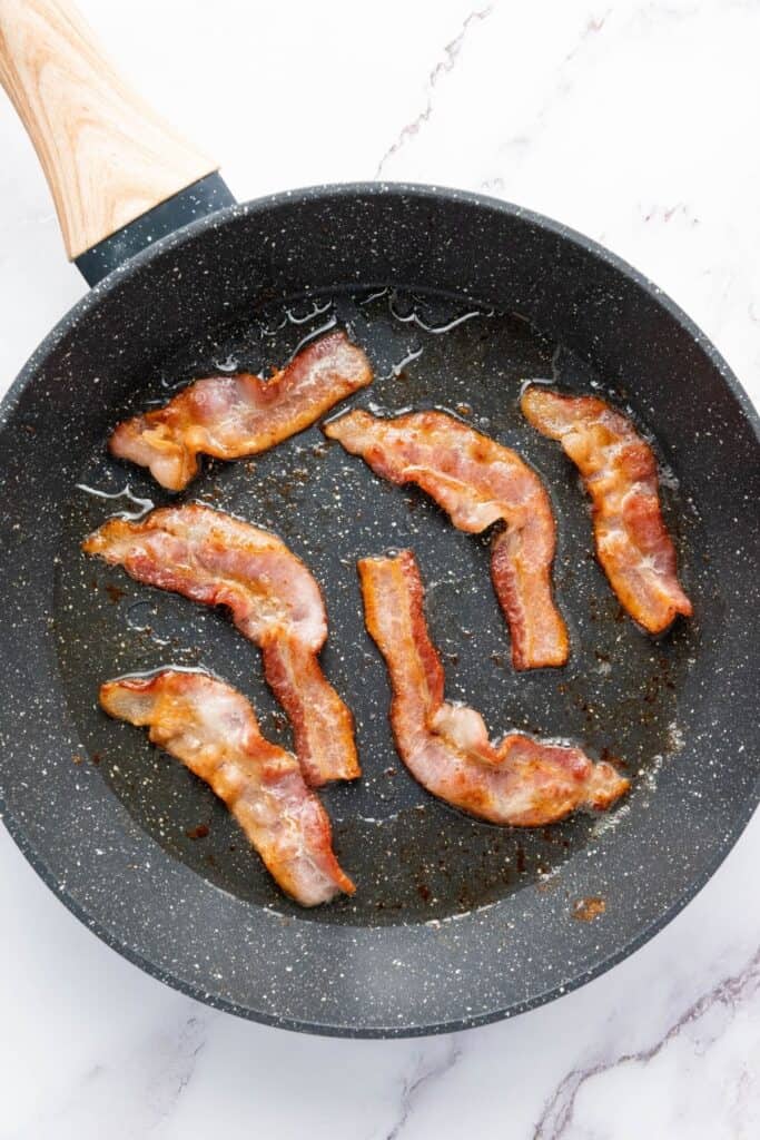 Six strips of bacon sizzling in a nonstick skillet with rendered fat, on a light marble countertop.