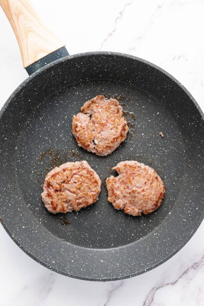 Three cooked sausage patties sit in a nonstick pan with a wooden handle on a light countertop.