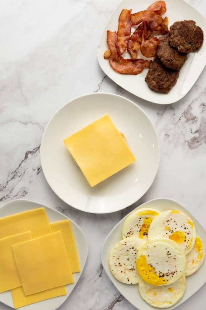 Four plates on marble: bacon and sausage, a cheese-topped sandwich, extra cheese slices, and cooked eggs.