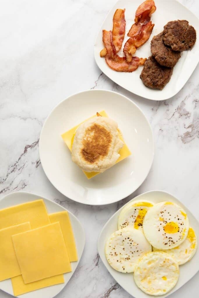 Four plates hold cooked bacon, sausage patties, cheese slices, fried eggs, and an English muffin breakfast sandwich.