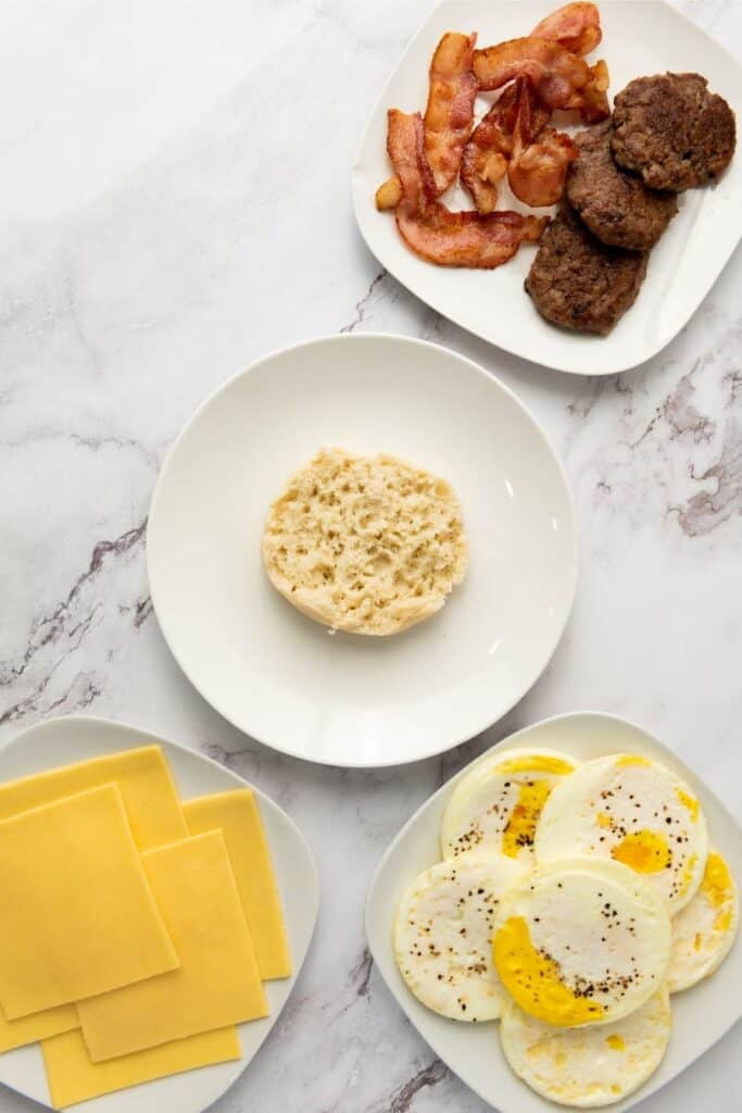 Four plates on marble display sliced cheddar, cooked eggs, an English muffin half, bacon, and sausage patties.