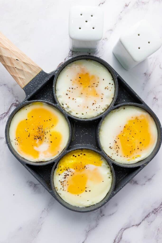 Four sunny-side-up eggs cook in a sectioned frying pan, sprinkled with salt and pepper; two white shakers behind.