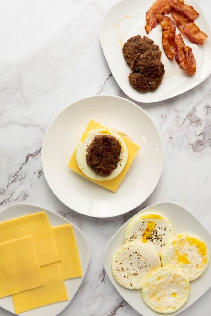 Cooked bacon, sausage patties, cheddar slices, and fried eggs on plates; one plate has a stacked sandwich.