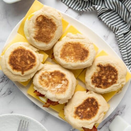 Six English muffin breakfast sandwiches neatly arranged on cheese slices, with eggs, forks, and a striped towel nearby.