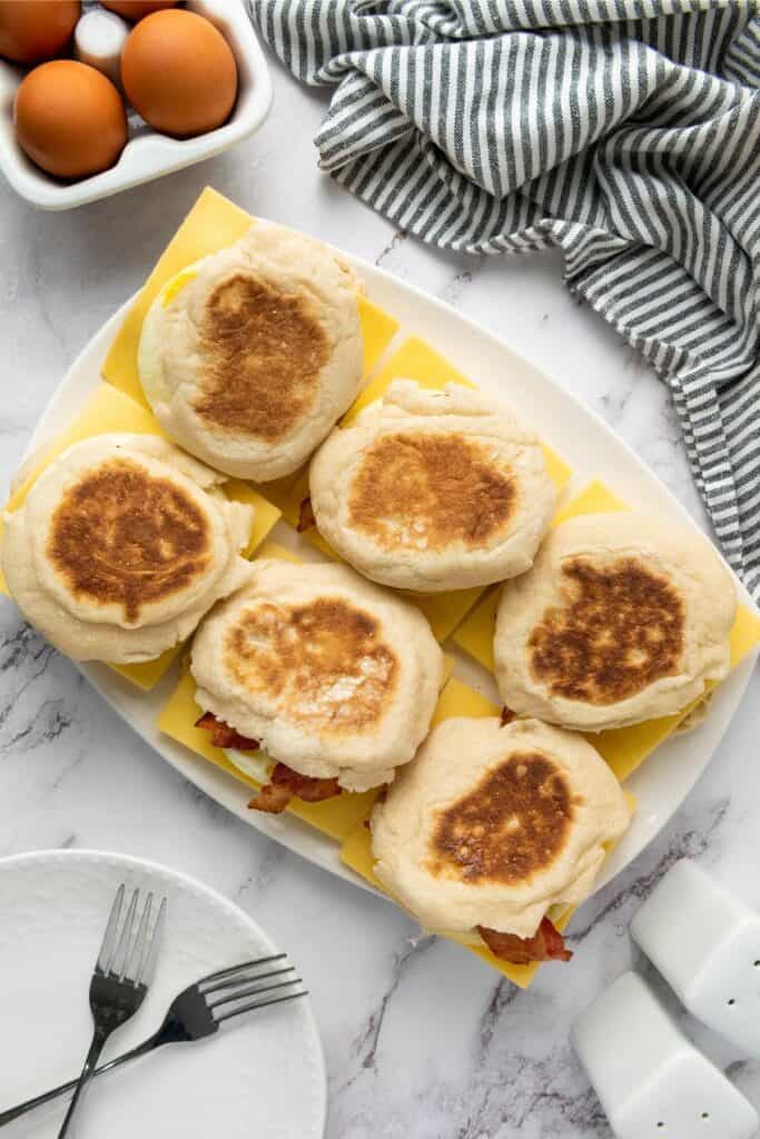 Six English muffin breakfast sandwiches neatly arranged on cheese slices, with eggs, forks, and a striped towel nearby.
