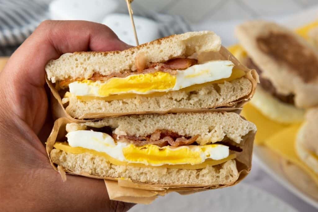 A hand holds half of a breakfast sandwich filled with egg, bacon, and melted cheese on an English muffin.