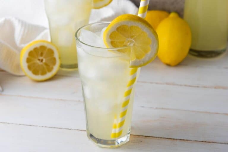 Iced lemonade in a glass with a yellow-striped straw and lemon slice, surrounded by whole and halved lemons.