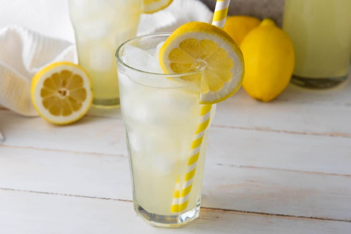 Iced lemonade in a glass with a yellow-striped straw and lemon slice, surrounded by whole and halved lemons.