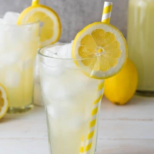 Iced lemonade in a glass with a striped straw and lemon slice, with extra lemons and lemonade behind.