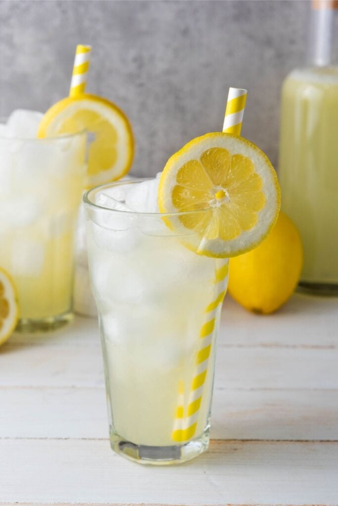 Iced lemonade in a glass with a striped straw and lemon slice, with extra lemons and lemonade behind.