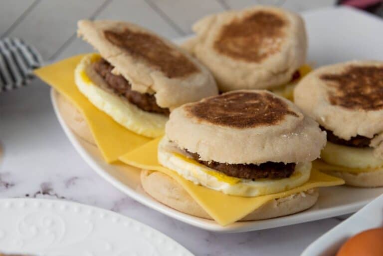 Breakfast sandwiches stacked on a white plate, made with English muffins, eggs, melted cheese, and sausage patties.