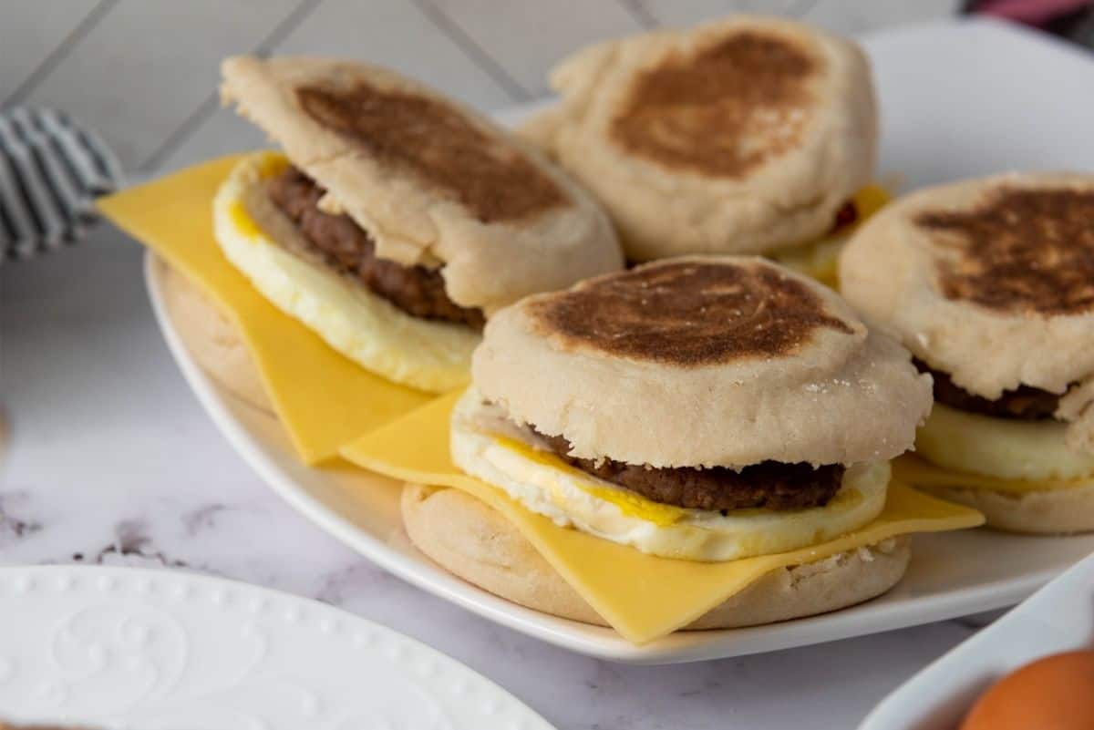 Breakfast sandwiches stacked on a white plate, made with English muffins, eggs, melted cheese, and sausage patties.