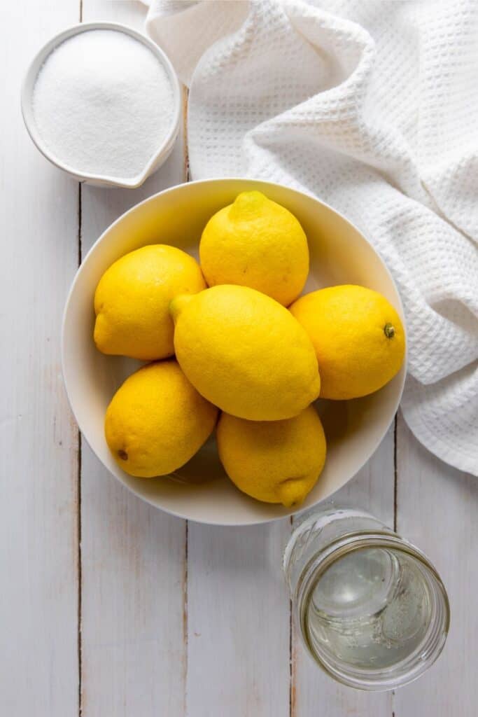 Six lemons in a white bowl sit on a white wooden surface beside a glass jar, sugar bowl, and textured cloth.