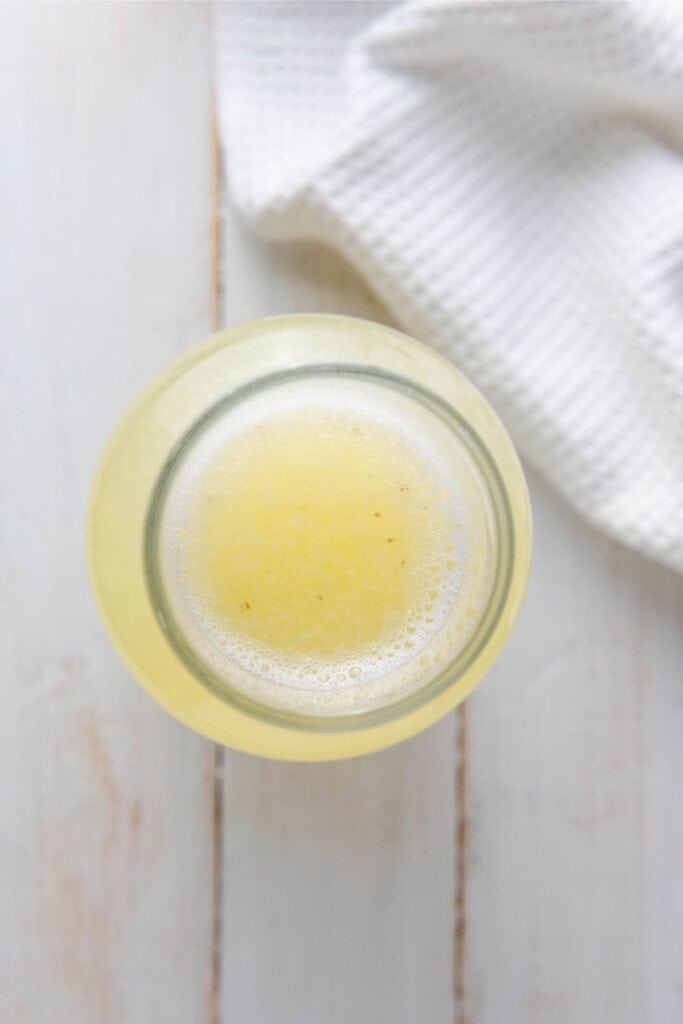 A top view of a glass of light yellow juice on a white wooden surface beside a textured white cloth.